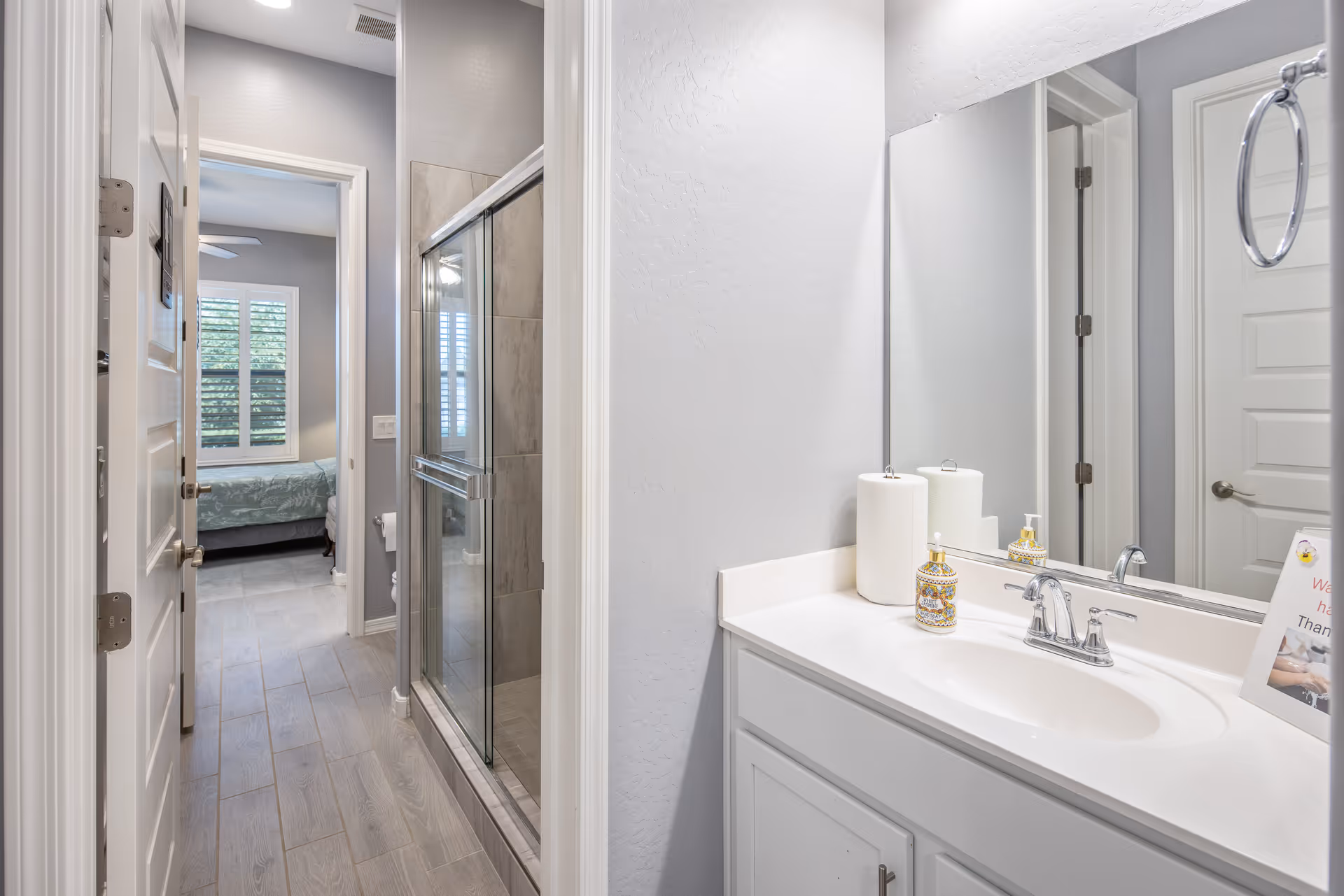 Bright bathroom with a white vanity and sink, mirror, glass-enclosed shower, and a view into an adjacent bedroom.