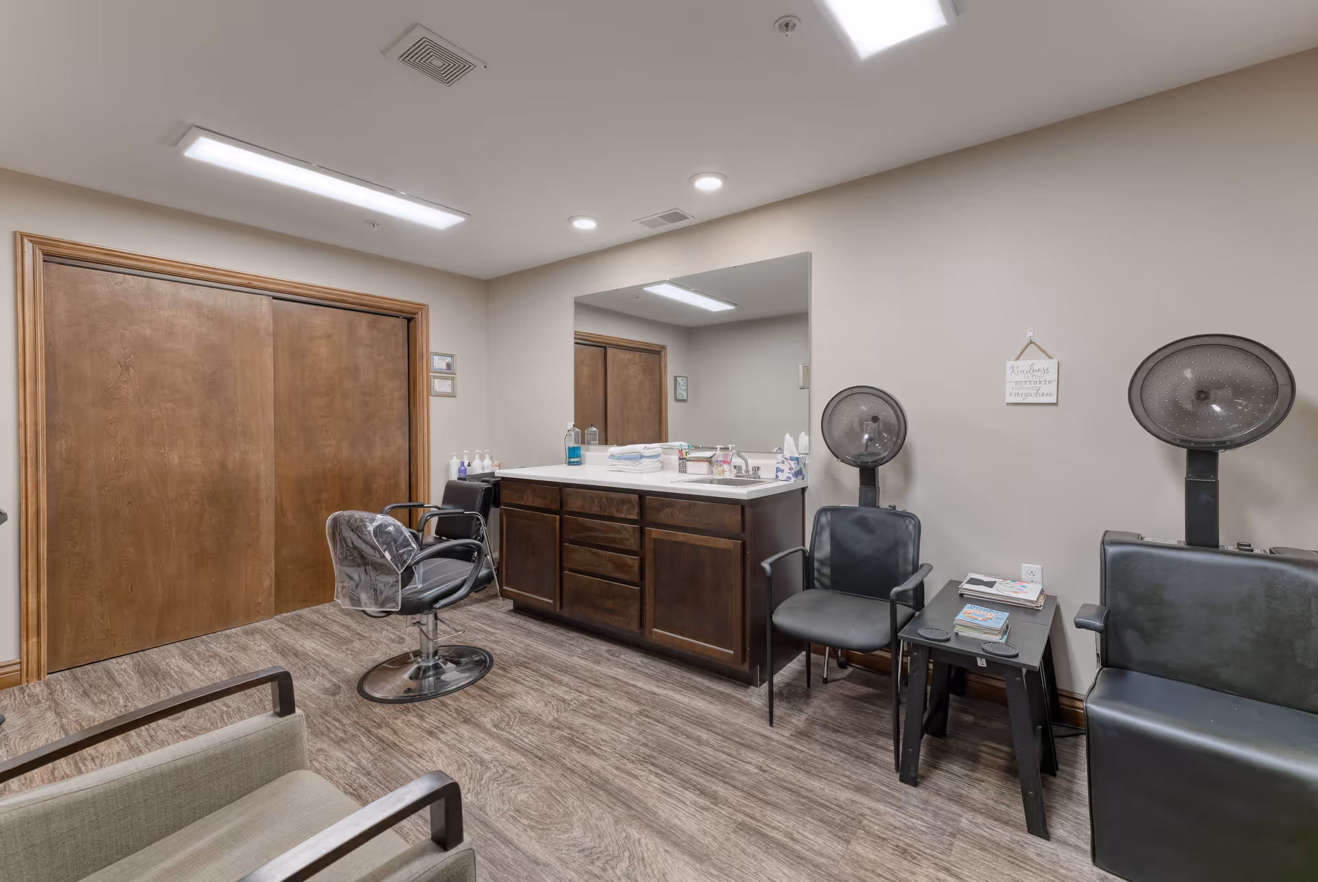 Interior view of a senior living facility's hair salon area with two black salon chairs under hair dryers, a black styling chair with a plastic cover, a large mirror above a wooden cabinet with a sink, and a small table with magazines. The room has wood flooring, beige walls, and a wooden sliding door.