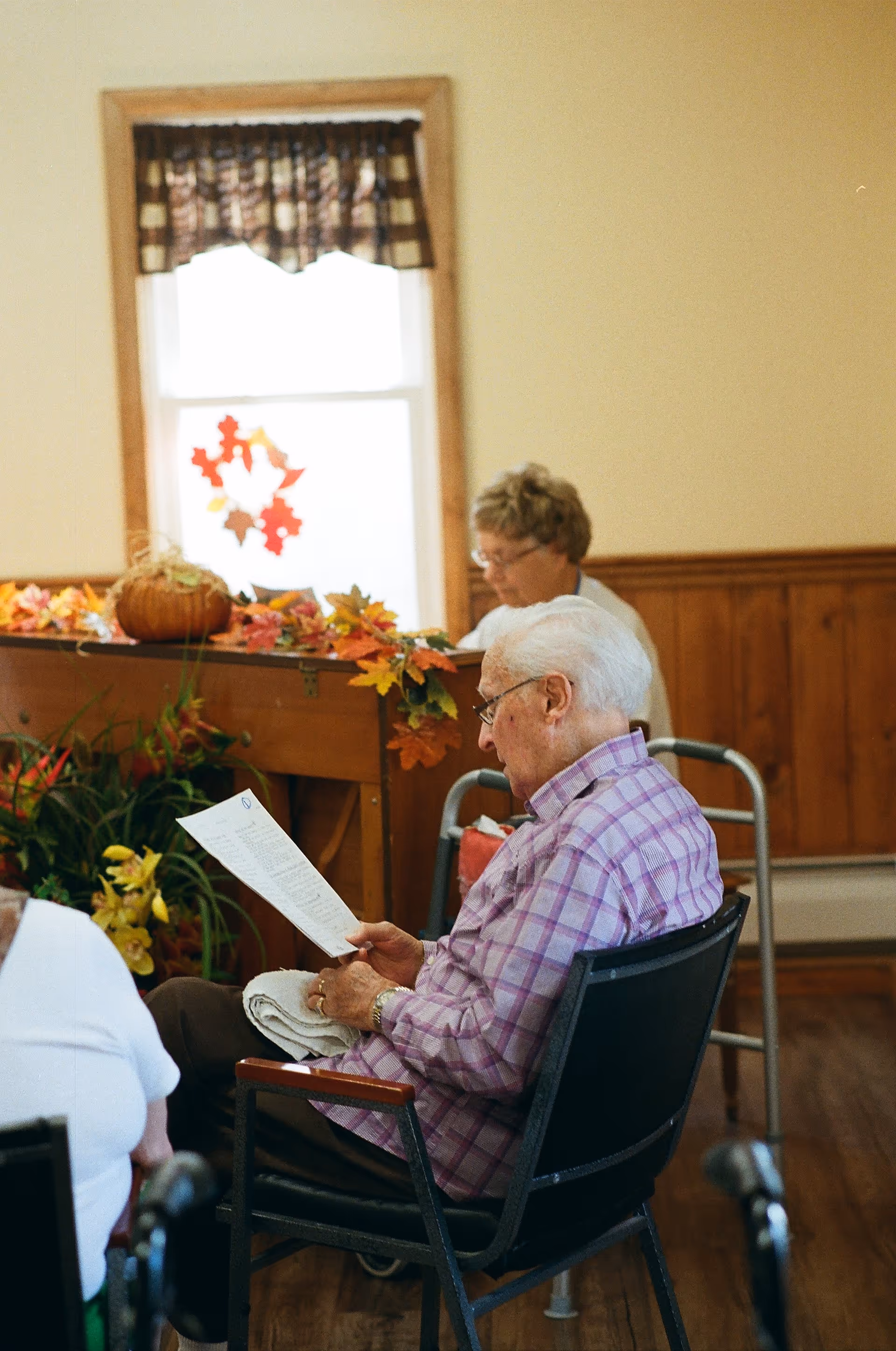 An elderly man with white hair and glasses sits in a chair reading a paper. Behind him, a woman is seated near a piano decorated with autumn leaves and a small pumpkin. The room has wooden paneling and a window with a checkered valance.