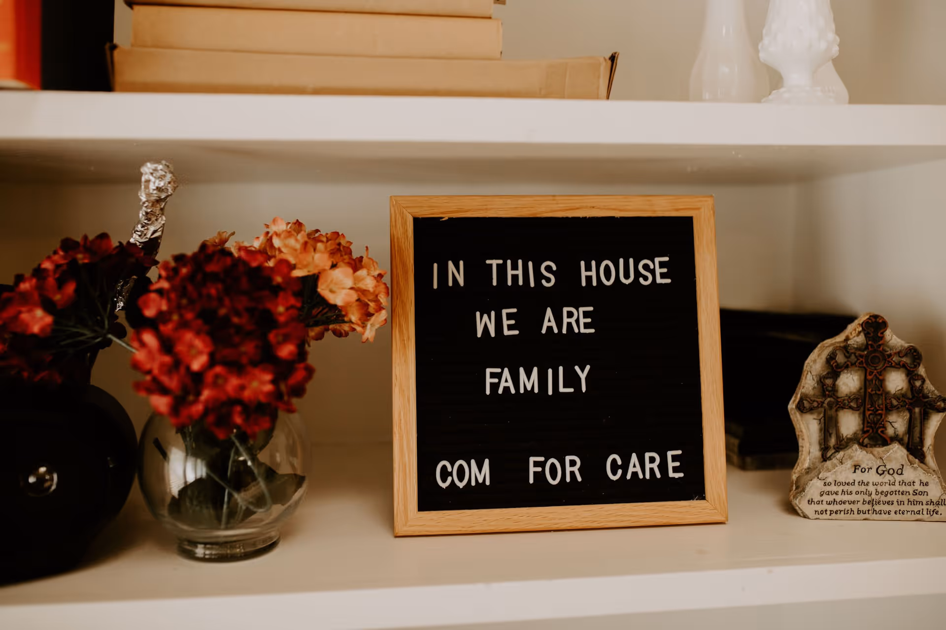 A shelf with a small vase of red and pink flowers, a framed letter board sign that reads 'IN THIS HOUSE WE ARE FAMILY COM FOR CARE', and a decorative religious plaque with a cross and a Bible verse.