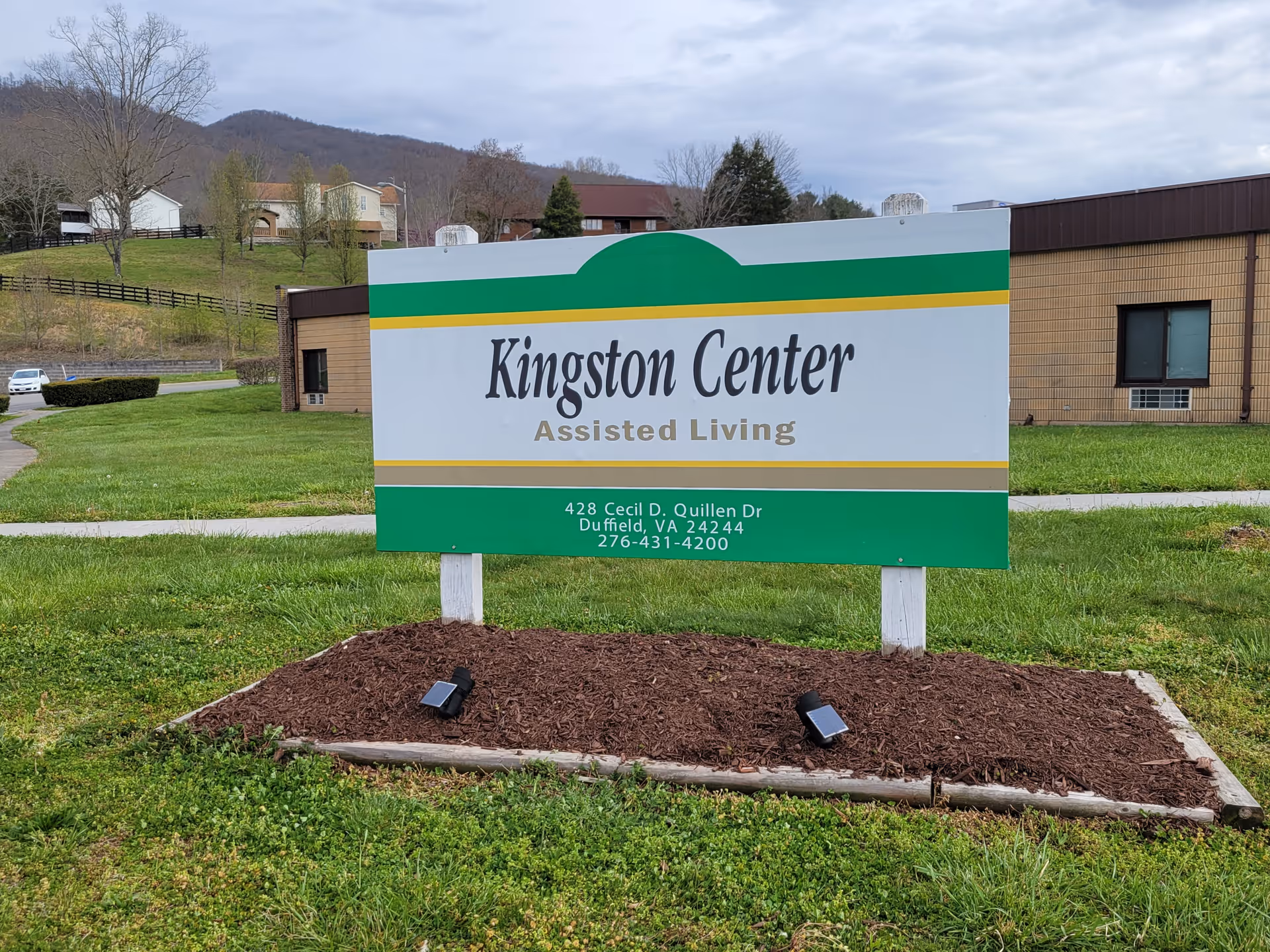 A freestanding sign reading "Kingston Center Assisted Living" on a grassy lawn in front of the facility building.