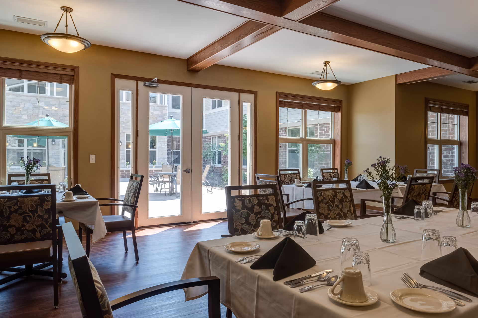 Sunlit dining room with tables set with white tablecloths, napkins, glassware and small floral centerpieces, and doors opening to an outdoor patio.