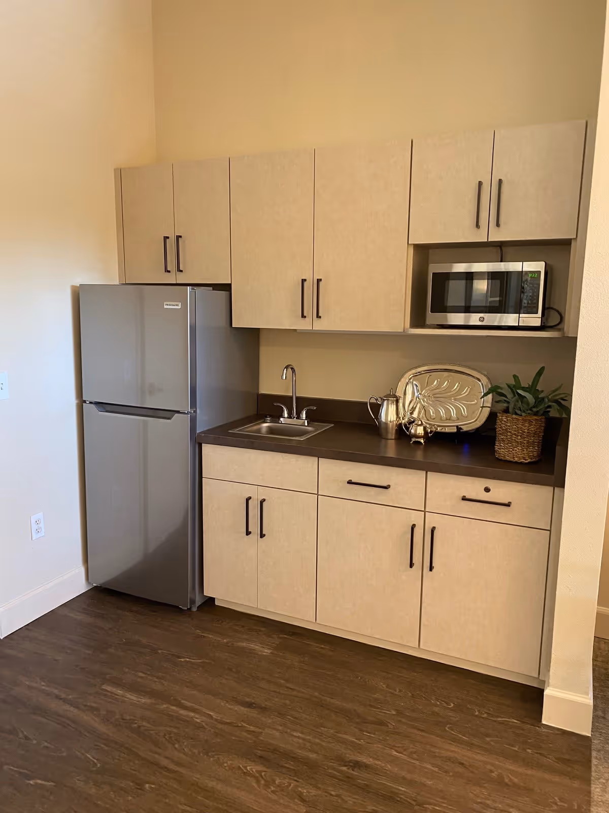 A small kitchen area with beige cabinets, a stainless steel refrigerator, a microwave, a sink, and a countertop with a silver tray, a silver pitcher, and a potted plant. The floor is wooden and the walls are light-colored.