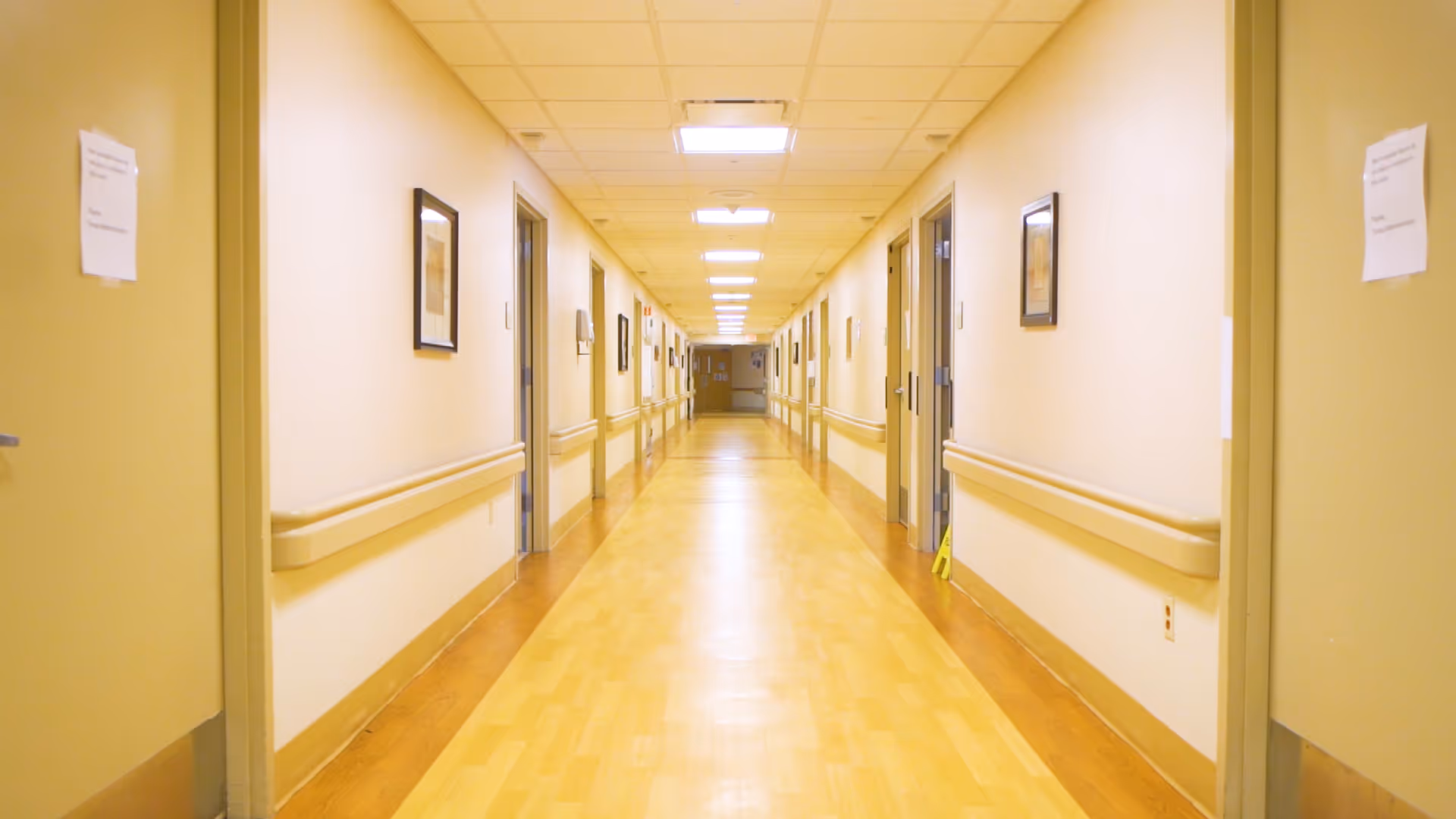 A long, well-lit hallway in a senior living facility with light-colored walls and wooden flooring. Doors line both sides of the corridor, each with handrails along the walls and framed pictures hanging. The ceiling has recessed lighting panels.