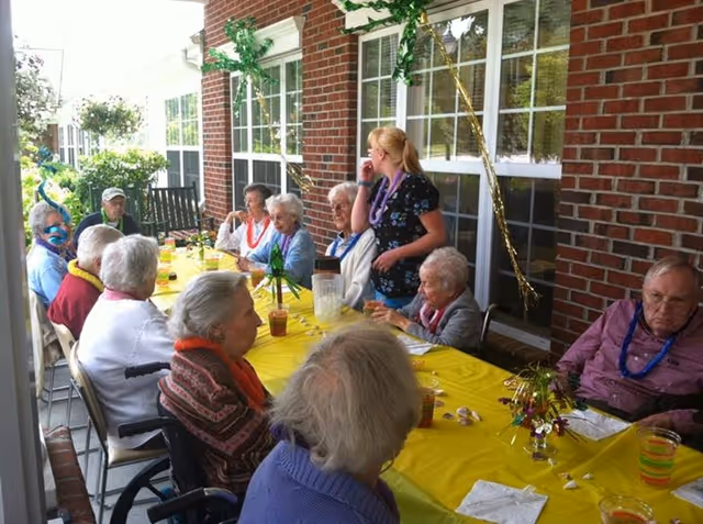 A group of elderly people sitting around a long table covered with a yellow tablecloth on a covered outdoor patio. A younger woman stands among them, engaging with the group. The table has drinks and small decorations, and the background shows a brick wall with windows and some greenery outside.