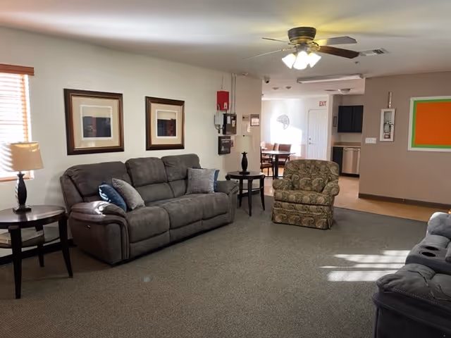 Communal living room with sofas, an armchair, side tables, a ceiling fan, and a view into a dining/kitchen area.