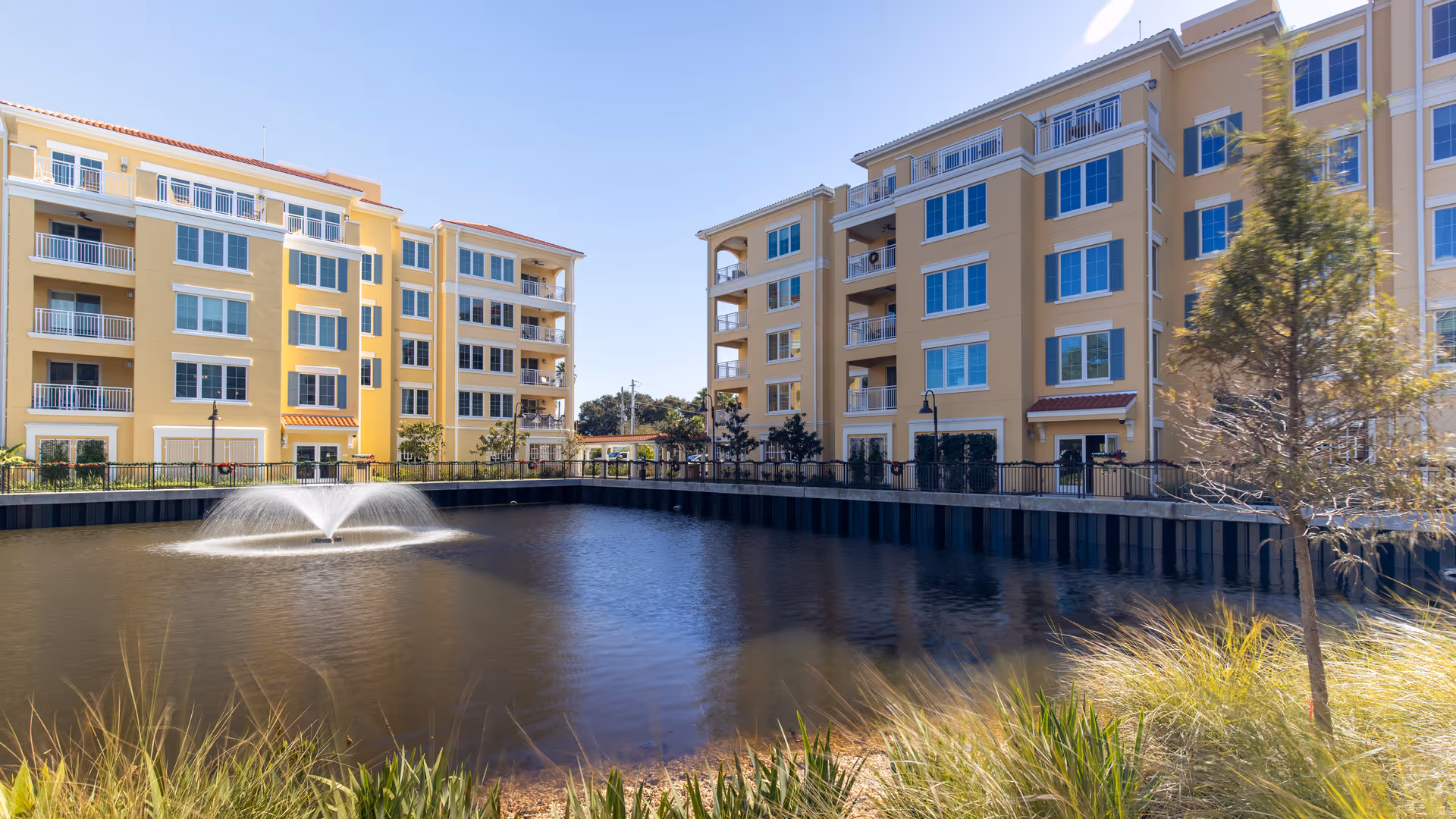 View of a pond with a water fountain in the center, surrounded by yellow multi-story residential buildings with balconies under a clear blue sky. There is greenery and a tree in the foreground near the water's edge.