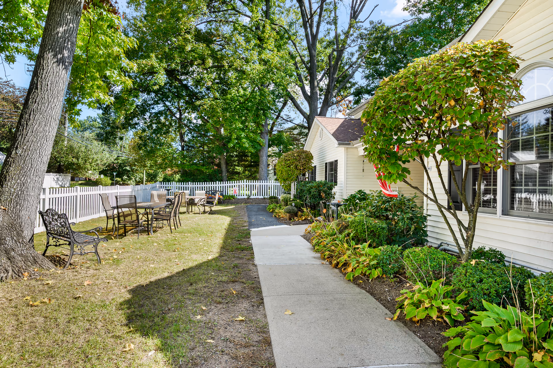 Outdoor garden area at Fox Trail Memory Care Living with a paved walkway, green trees, bushes, and a white picket fence. There are several metal chairs and tables arranged on the grass near the fence, and a bench is placed under a large tree. The side of a light-colored building with windows and an American flag is visible on the right.