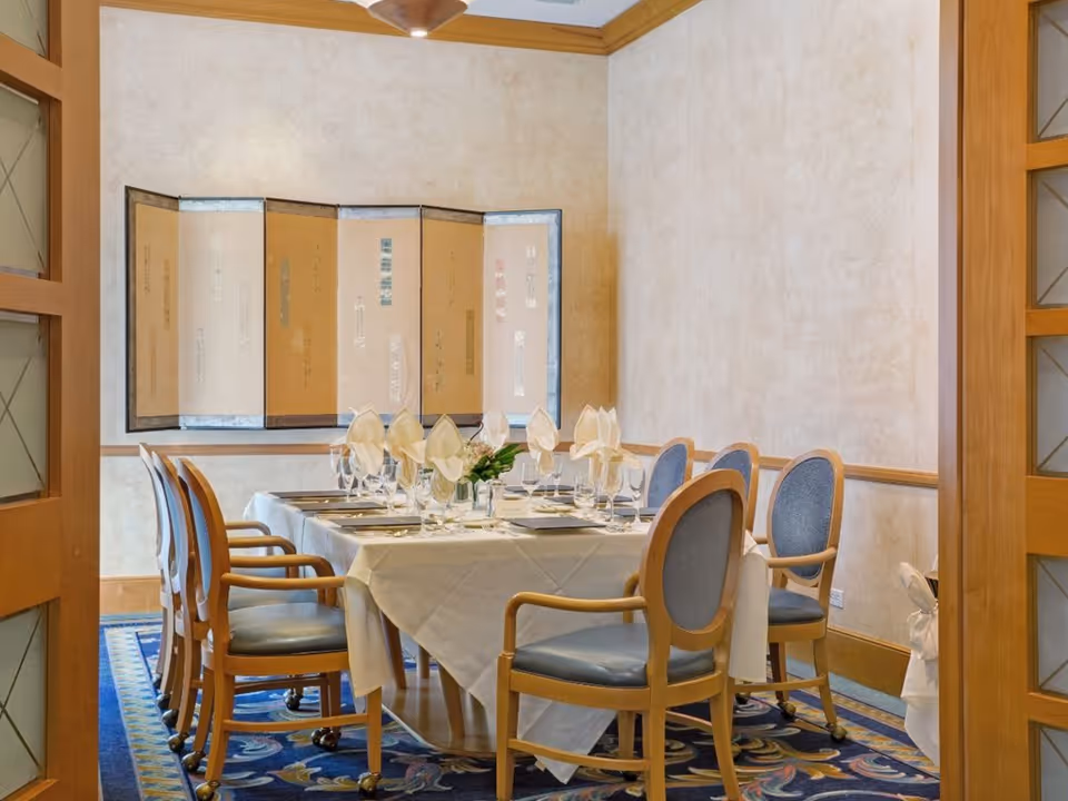 A formal dining room with a rectangular table covered with a white tablecloth, set with glassware, plates, and folded napkins. The room has wooden framed chairs with blue cushions, a decorative wall panel, and a patterned blue carpet.