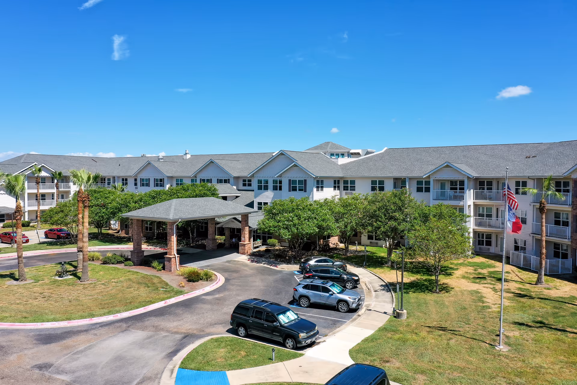 Exterior view of Solstice Senior Living at Corpus Christi, showing a large multi-story building with a covered entrance, several parked cars, palm trees, and a flagpole with the American and Texas flags against a clear blue sky.