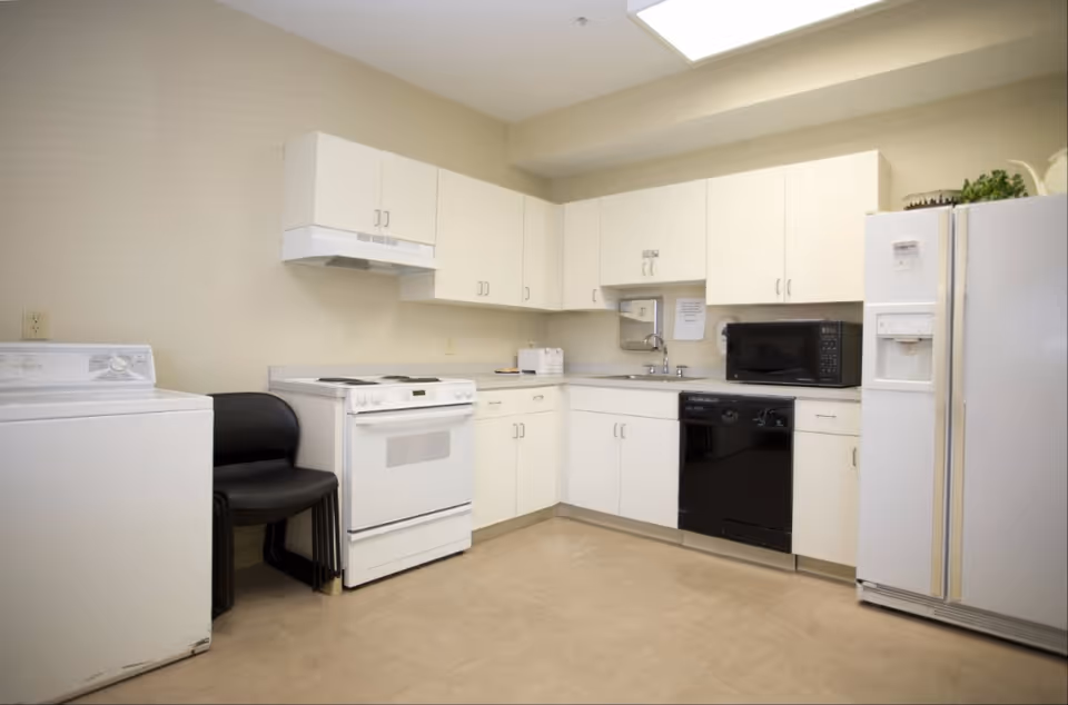 A clean kitchen area with white cabinets, a white stove, a black dishwasher, a microwave, a white refrigerator with an ice dispenser, and a washing machine. There are two black chairs stacked next to the washing machine. The walls are beige and the floor is light-colored.
