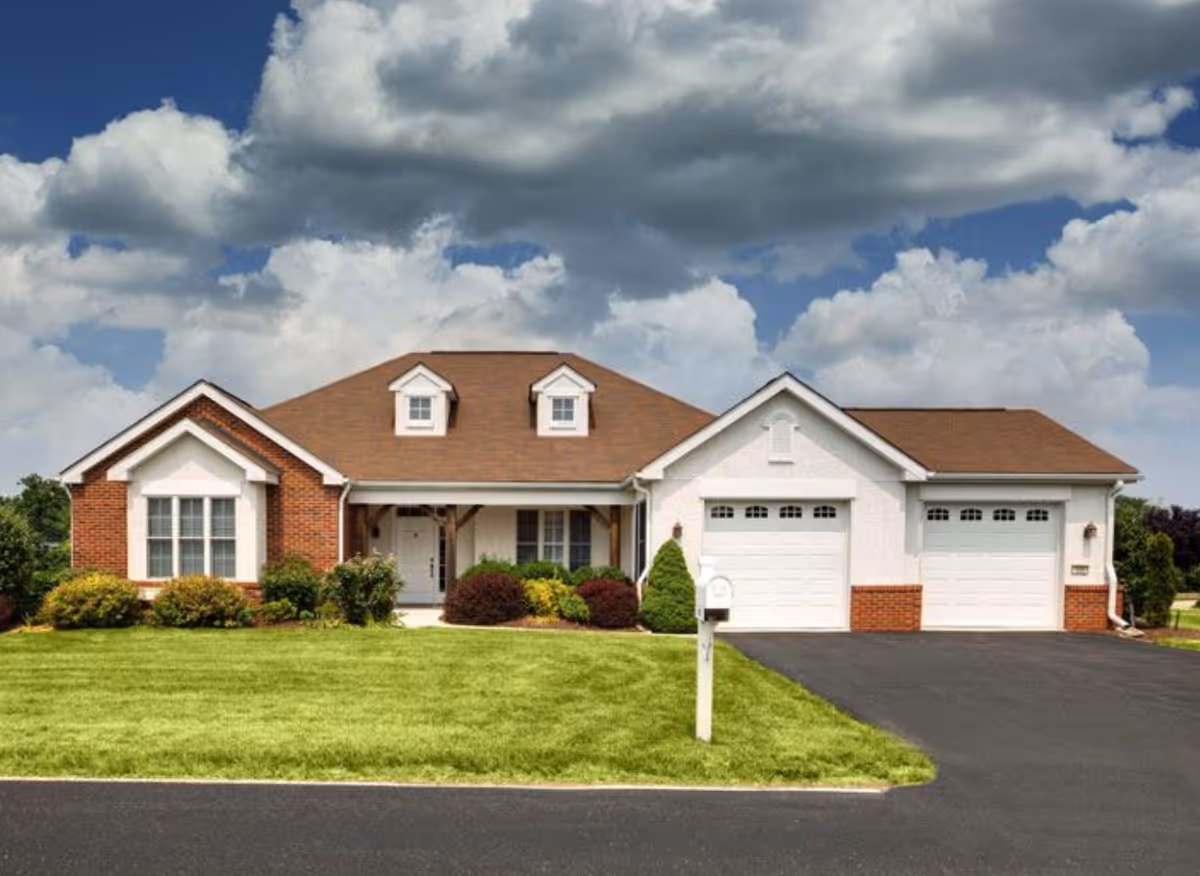 Single-story residential building with a brown roof, white and brick exterior walls, two dormer windows, a front porch, a well-maintained lawn, and a driveway leading to a double garage under a partly cloudy sky.
