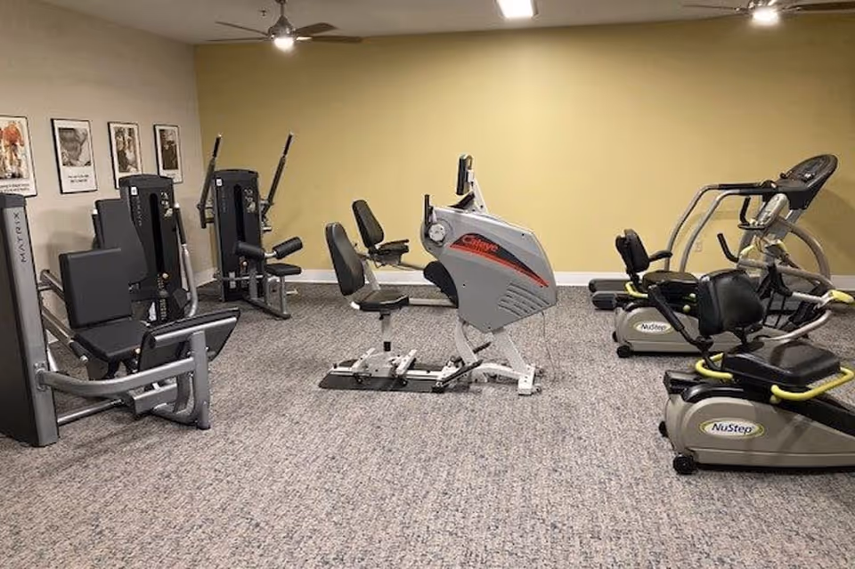 A fitness room with various exercise equipment including seated leg press machines, recumbent bikes, and elliptical machines. The room has beige walls, carpeted flooring, and ceiling fans with lights. There are framed pictures on one wall.