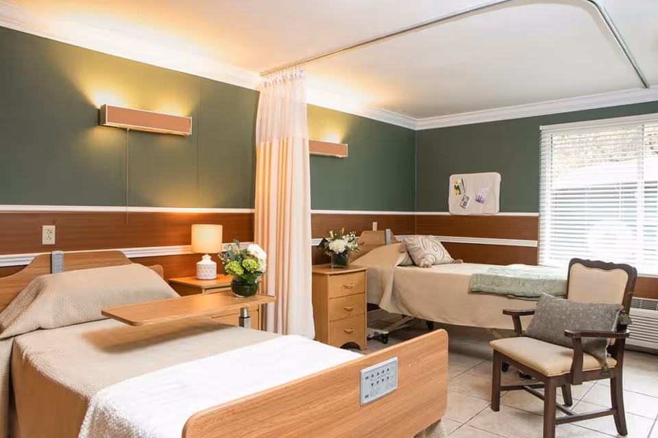 A shared senior living room with two beds, a bedside table, and a chair, featuring warm lighting and green walls.