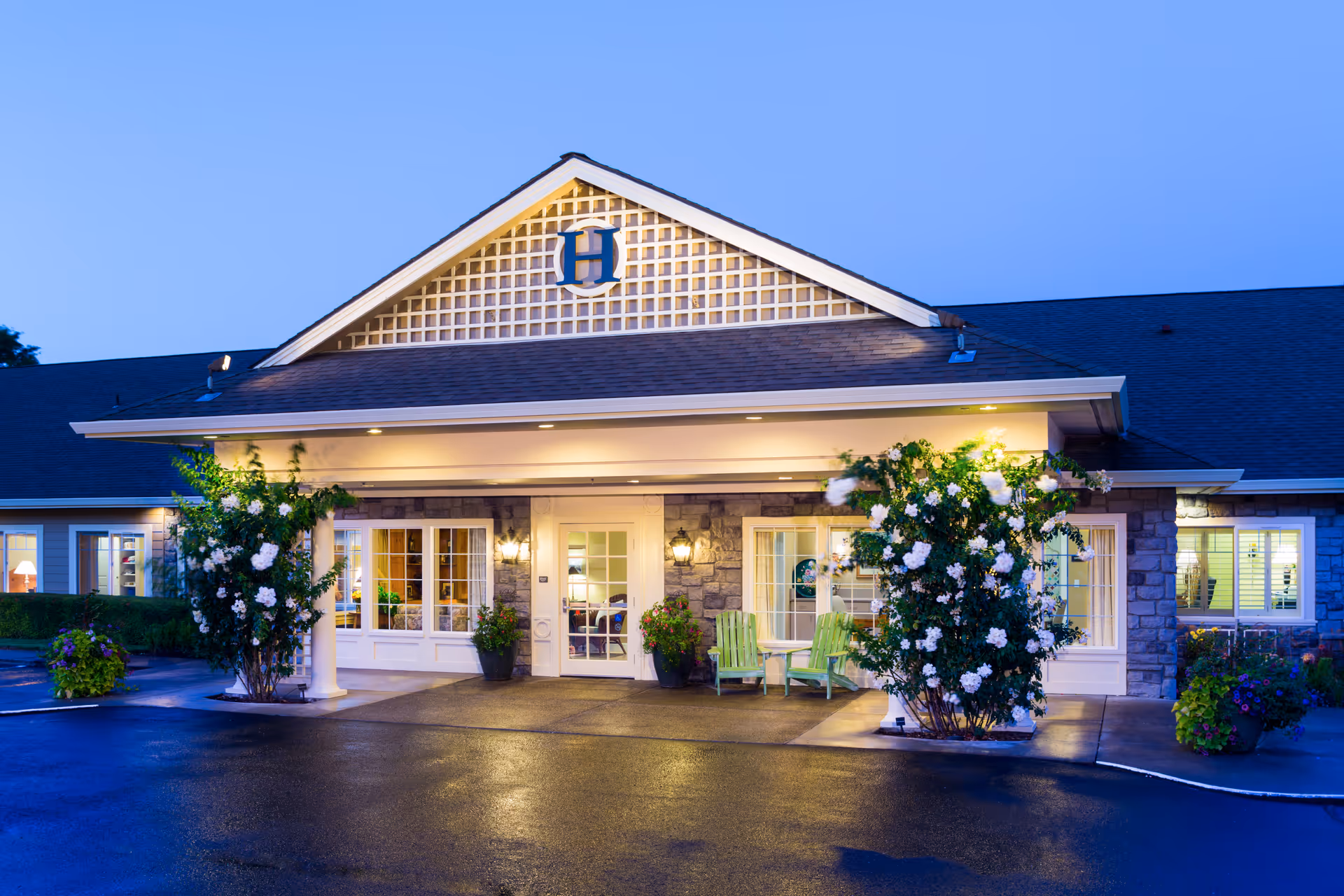 Exterior front entrance of The Hampton and Ashley Inn Memory Care Community at dusk, featuring a covered porch with white columns, two green chairs, flowering bushes on either side, and warm lights illuminating the building.