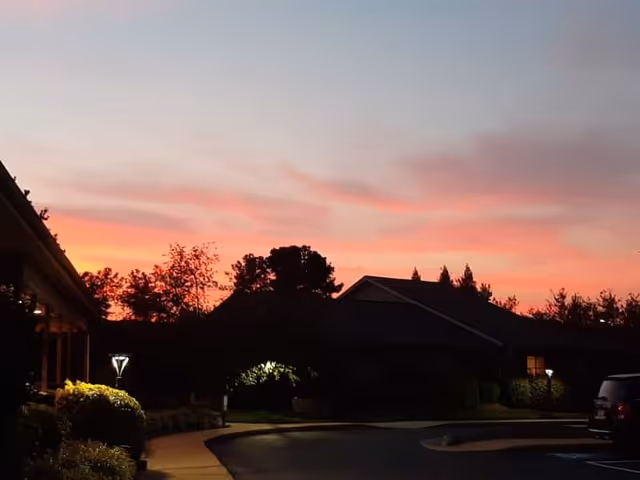 A twilight view of a senior living facility with a colorful sunset sky in the background. The scene shows a curved driveway, illuminated street lamps, bushes, trees, and the silhouette of buildings with some windows lit.