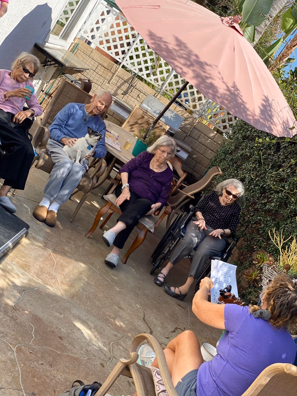 Four elderly women sitting outdoors on chairs under a large pink patio umbrella. One woman is in a wheelchair, another is holding a small dog on her lap. A person with short hair wearing a purple shirt is sitting on a chair facing them, holding a sheet of paper and a musical instrument, possibly leading an activity or singing session. The setting is a patio area with a lattice fence and greenery in the background.