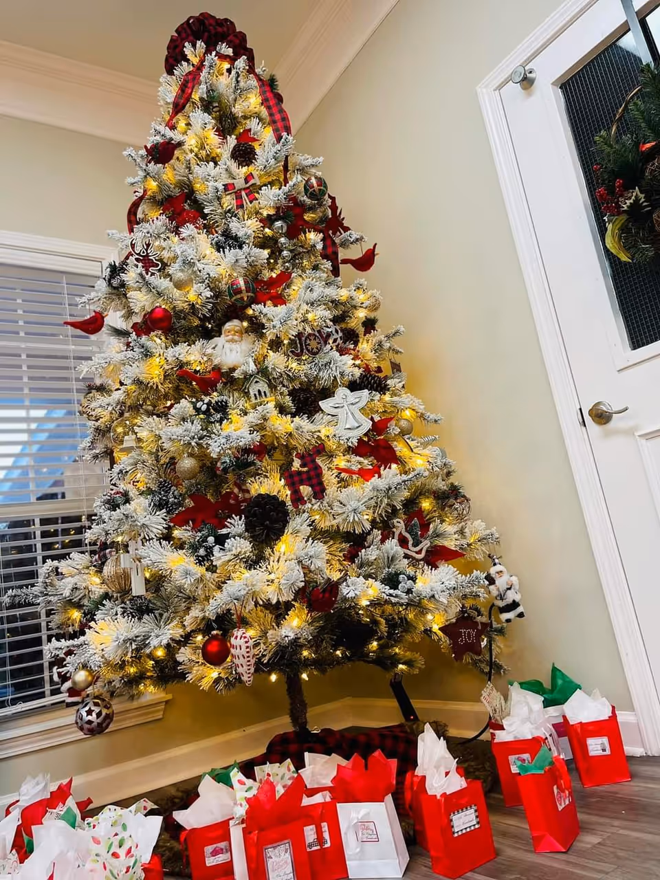 A decorated Christmas tree with white frosted branches, red and gold ornaments, pine cones, and lights stands in the corner of a room near a window and a white door with a holiday wreath. Several red and white gift bags with tissue paper are arranged on the floor around the base of the tree.