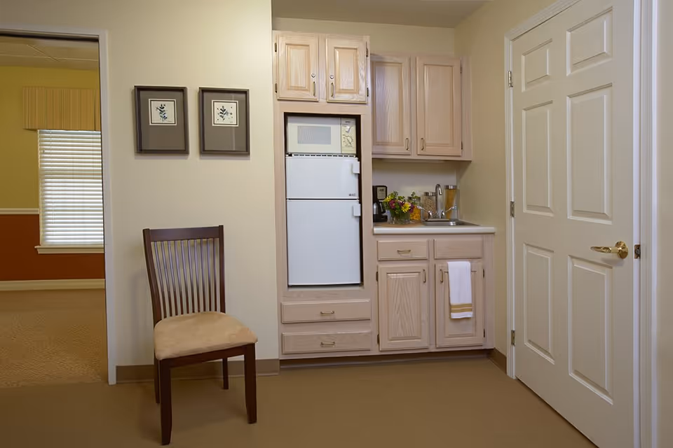 Small kitchenette area with light wood cabinets, a white mini refrigerator, and a microwave above it. A sink with a faucet is on the right side of the counter, with a white towel hanging from the cabinet handle. A wooden chair with a beige cushion is placed against the wall next to two framed pictures. A doorway to another room with a window and blinds is visible on the left, and a closed white door with a gold handle is on the right.