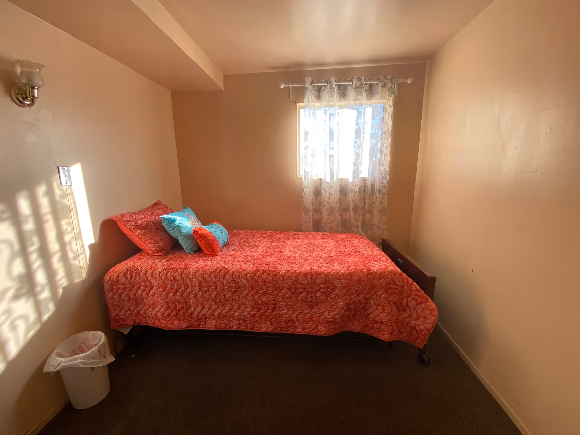 A small bedroom with a single bed covered in a red patterned bedspread and several pillows in red and blue. There is a window with sheer patterned curtains letting in sunlight, beige walls, a wall-mounted light fixture, and a white trash bin on the floor near the bed.