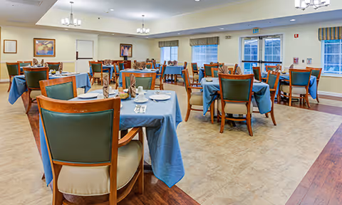 A spacious dining room with multiple tables covered in blue tablecloths, each set with plates, cups, and silverware. The room has wooden chairs with green cushions, large windows with striped valances, and framed artwork on the walls. Ceiling lights provide bright illumination.
