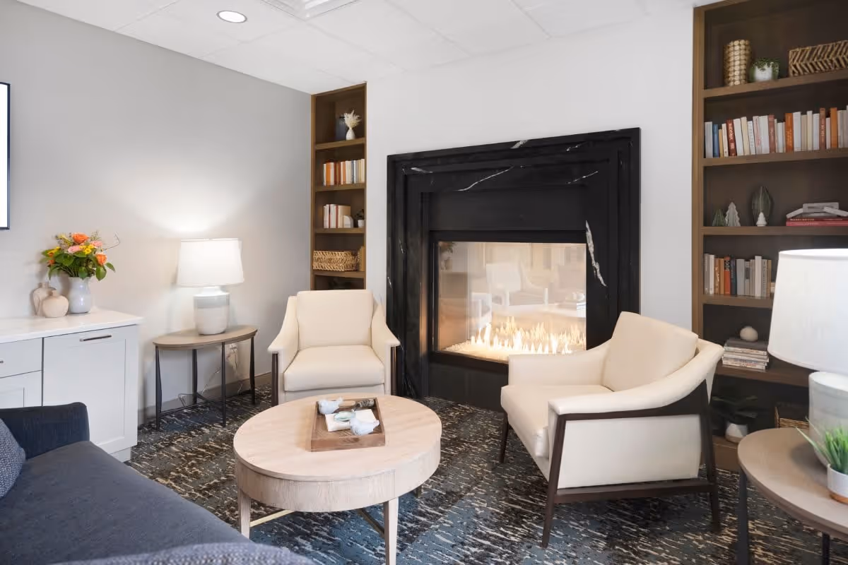 A cozy living room area with a modern black fireplace in the center, two cream-colored armchairs, a round wooden coffee table with a tray, a dark sofa, built-in wooden bookshelves filled with books and decorative items, a side table with a lamp, and a cabinet with a vase of flowers.