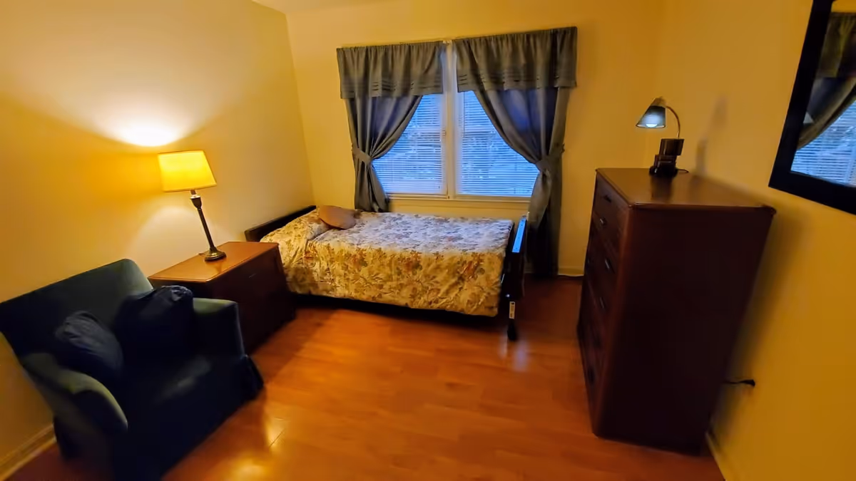 Small furnished bedroom with a single bed under a curtained window, a dresser, armchair and bedside lamp.