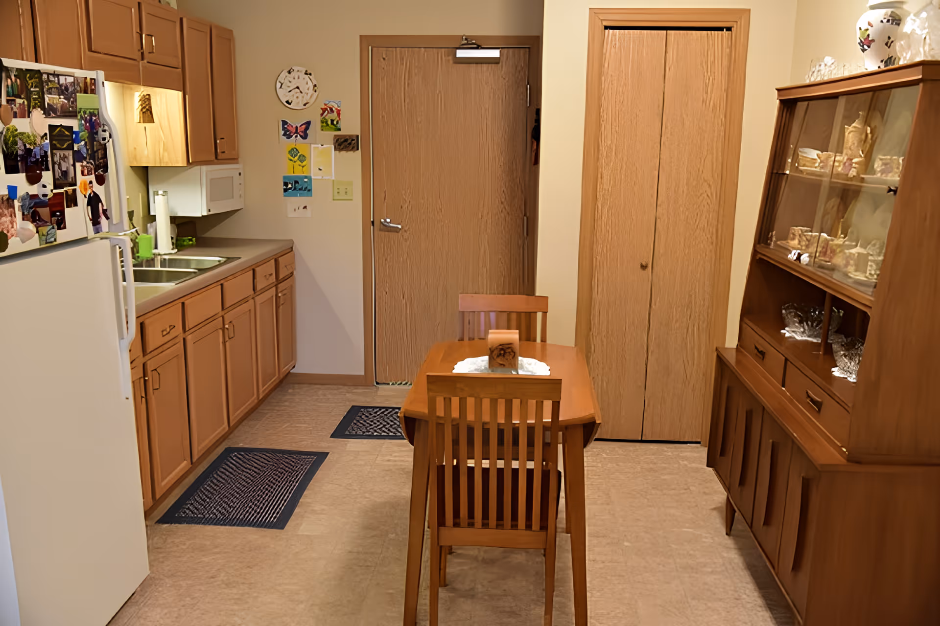 A small kitchen and dining area with wooden cabinets, a white refrigerator covered with photos and magnets, a double sink, a microwave, and a wooden dining table with two chairs. There is a wooden cabinet with glass doors displaying dishes and glassware. The walls have some colorful artwork and a clock. Two closed wooden doors are visible in the background.