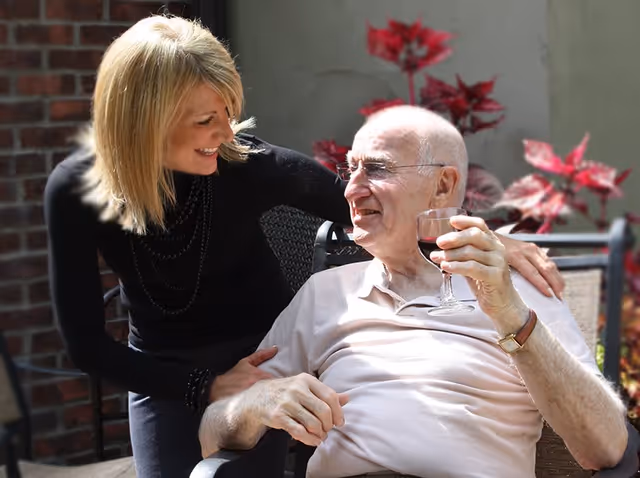 An elderly man sitting outdoors in a chair holding a glass of red wine, smiling and looking at a woman who is leaning towards him with a warm smile. There are red-leaved plants and a brick wall in the background.
