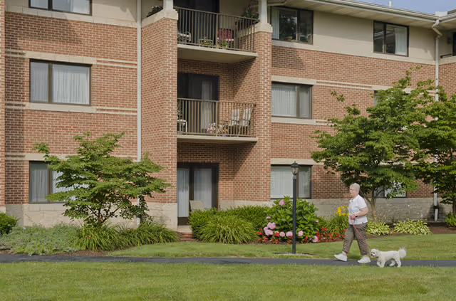 A senior woman walking a small white dog on a leash along a paved path in front of a three-story brick apartment building with balconies and windows. The area is landscaped with green grass, bushes, and flowering plants.
