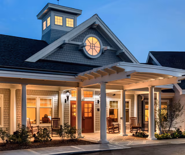 Well-lit building entrance with a covered porch, white columns and rocking chairs at dusk.