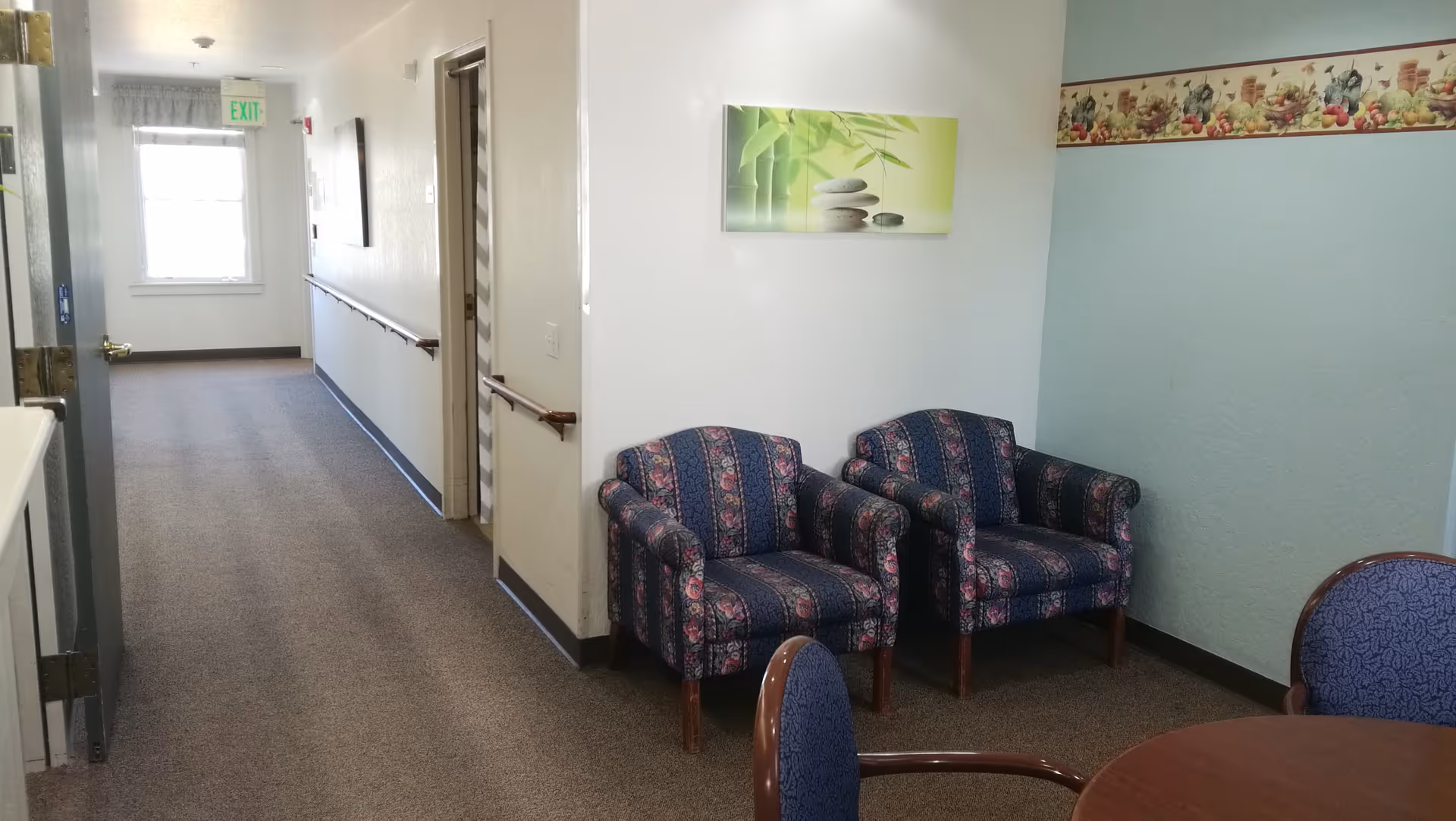 A hallway in a senior living facility with two patterned armchairs against a wall, a round wooden table with chairs in the foreground, a framed picture of bamboo and stones on the wall, and a decorative border with fruit motifs near the ceiling. The hallway has a window at the end with an exit sign above it and handrails along the walls.