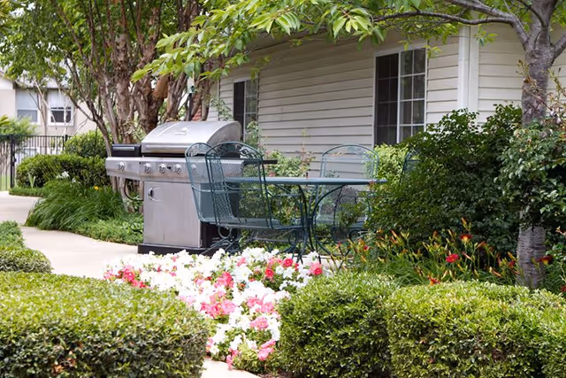 Outdoor patio area at DaySpring Senior Living featuring a metal table with four chairs, a stainless steel barbecue grill, surrounded by green bushes, flowering plants, and trees next to a beige building with windows.