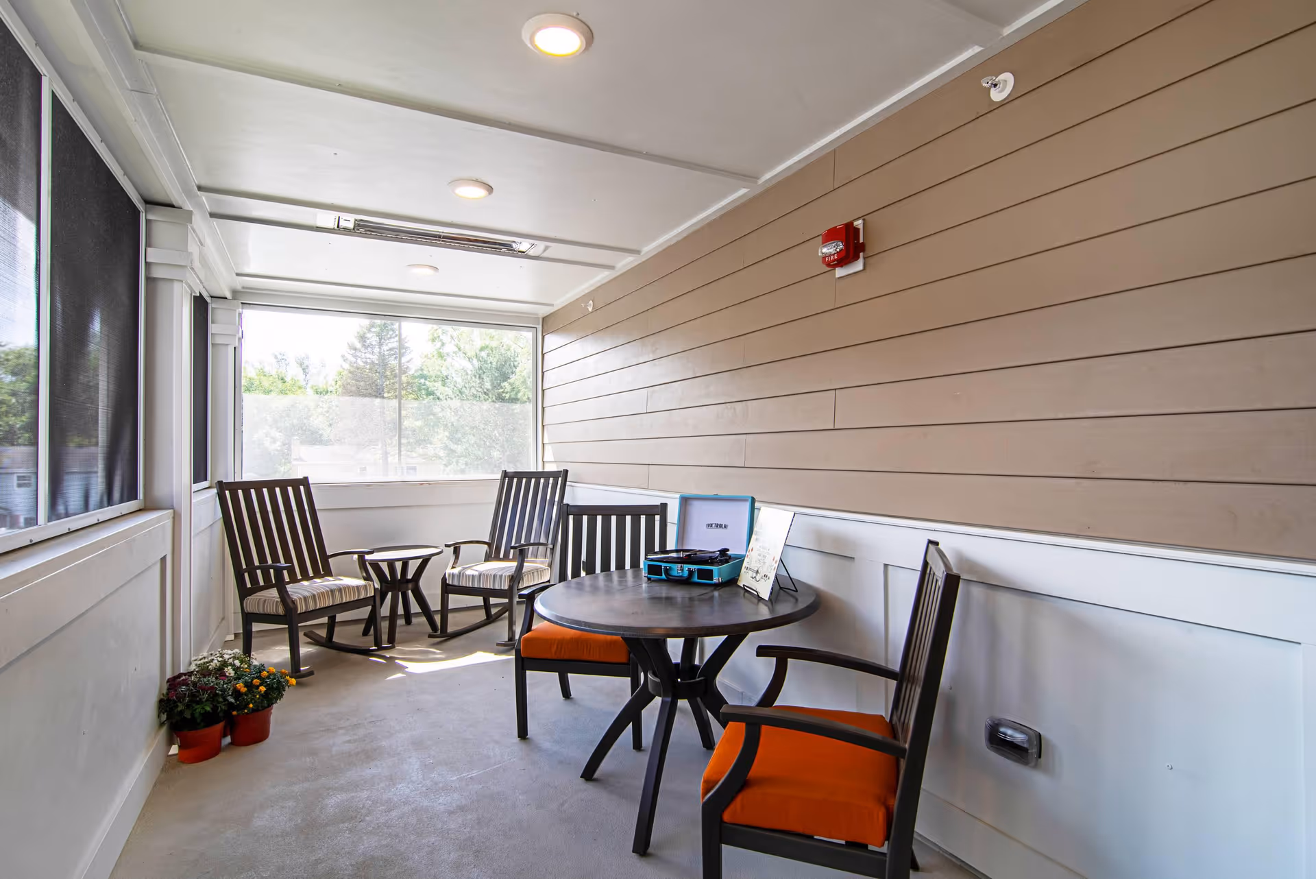 A bright enclosed porch area with beige siding and white trim, featuring a round table with two chairs that have orange cushions, two wooden armchairs with striped cushions, a small side table, and potted flowers on the floor near the window. A blue record player and a framed picture are on the round table.