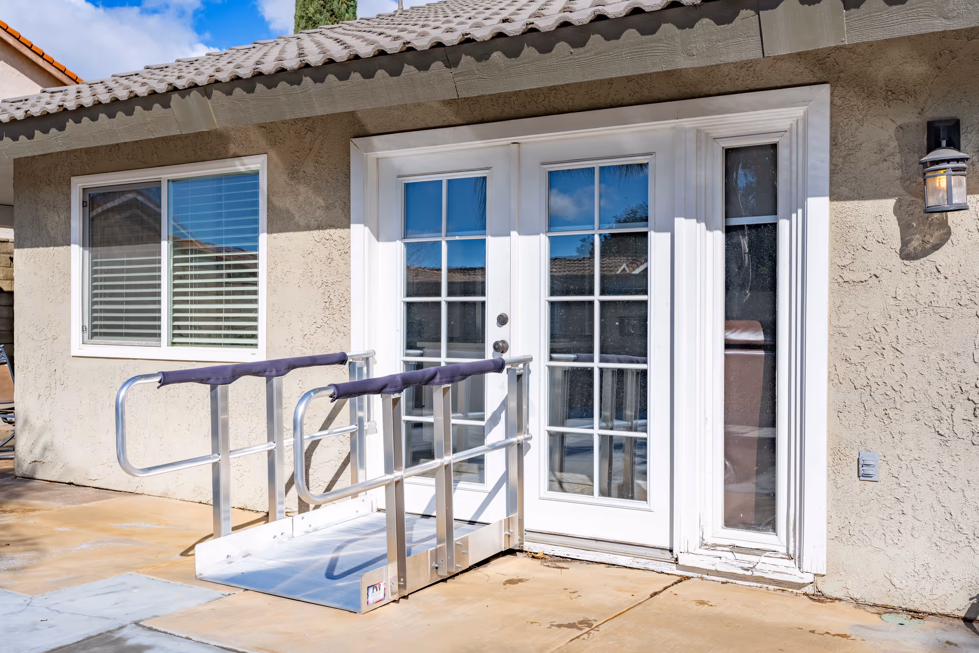 Exterior view of a building wall with a window and a set of white French doors. A metal wheelchair ramp with padded handrails leads up to the doors. The wall is beige stucco with a small outdoor light fixture mounted on the right side.