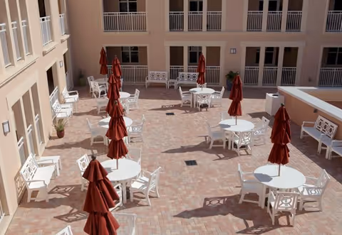 Outdoor courtyard area with multiple white round tables and chairs arranged on a brick-paved floor. Each table has a closed red umbrella. The courtyard is surrounded by a beige building with windows and balconies.