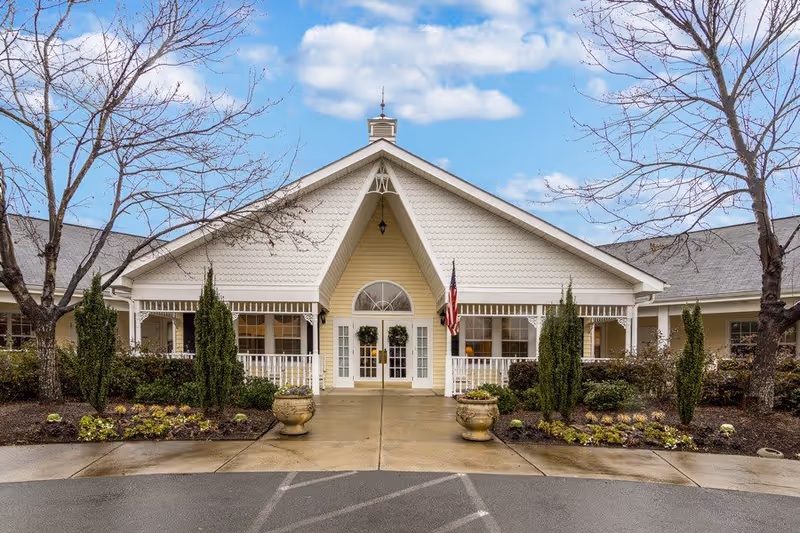 Front exterior view of TerraBella Hillsborough facility showing a large entrance with white double doors, a peaked roof with decorative trim, two potted plants on either side of the walkway, landscaped bushes and trees, and an American flag mounted near the entrance under a partly cloudy sky.