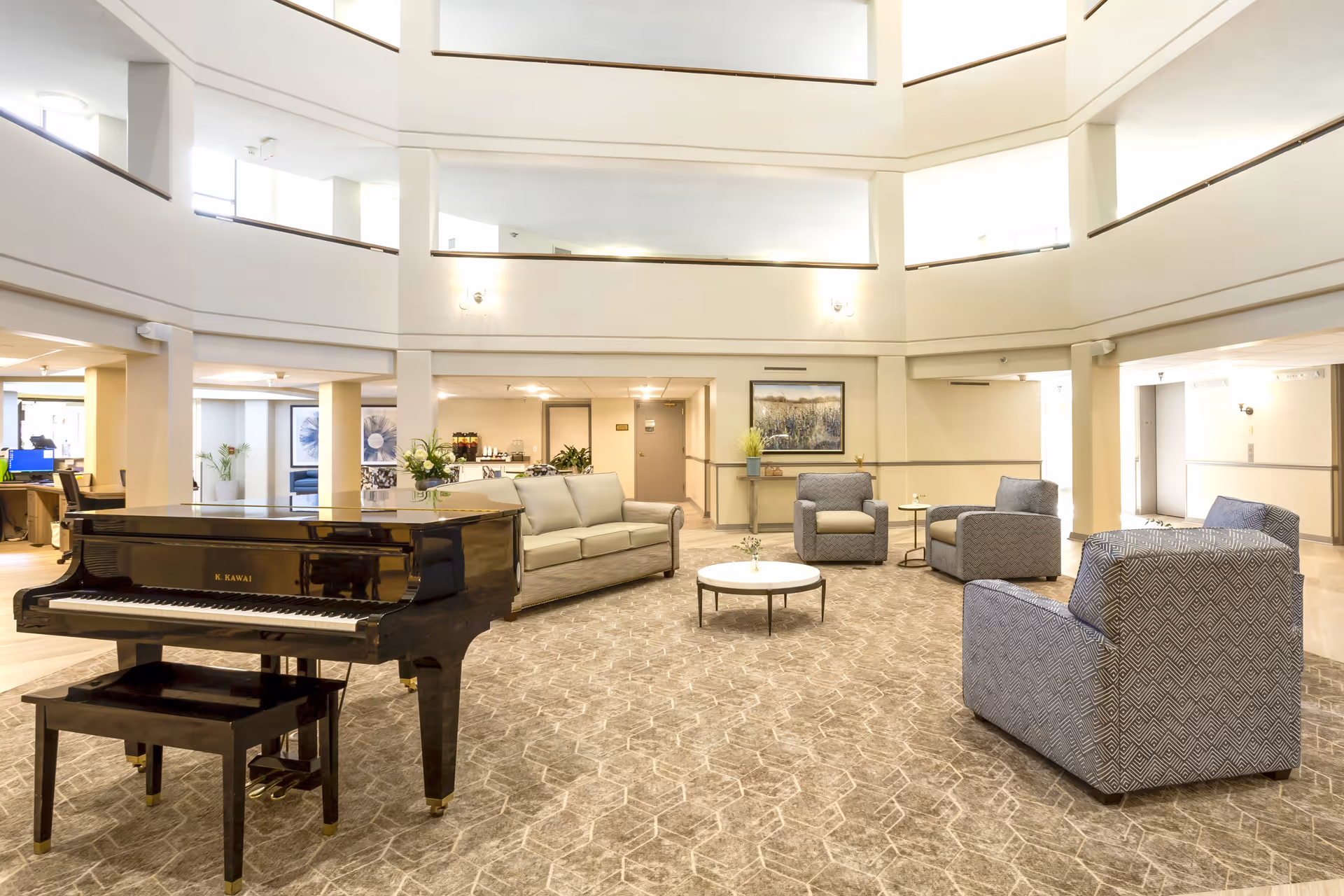 A spacious senior living facility common area with a grand piano, several armchairs, a sofa, and a round coffee table on a patterned carpet. The room has high ceilings with multiple levels visible and soft lighting on the walls. There are plants and framed artwork decorating the space.