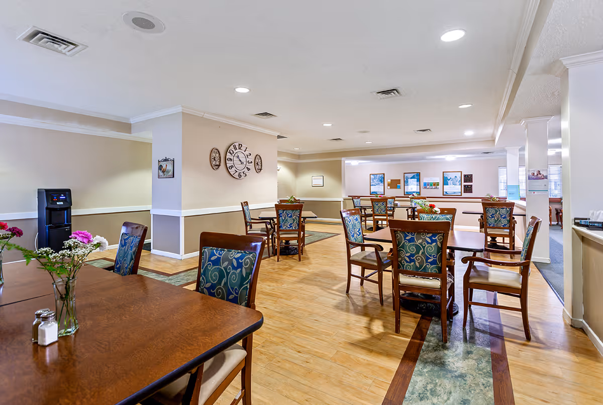 A spacious dining room in a senior living facility with multiple wooden tables and chairs featuring patterned upholstery. The room has light-colored walls with a large clock and decorative wall hangings. There are flowers in vases on the tables and a water dispenser in the corner. The floor is a mix of wood and patterned tiles, and the ceiling has recessed lighting.