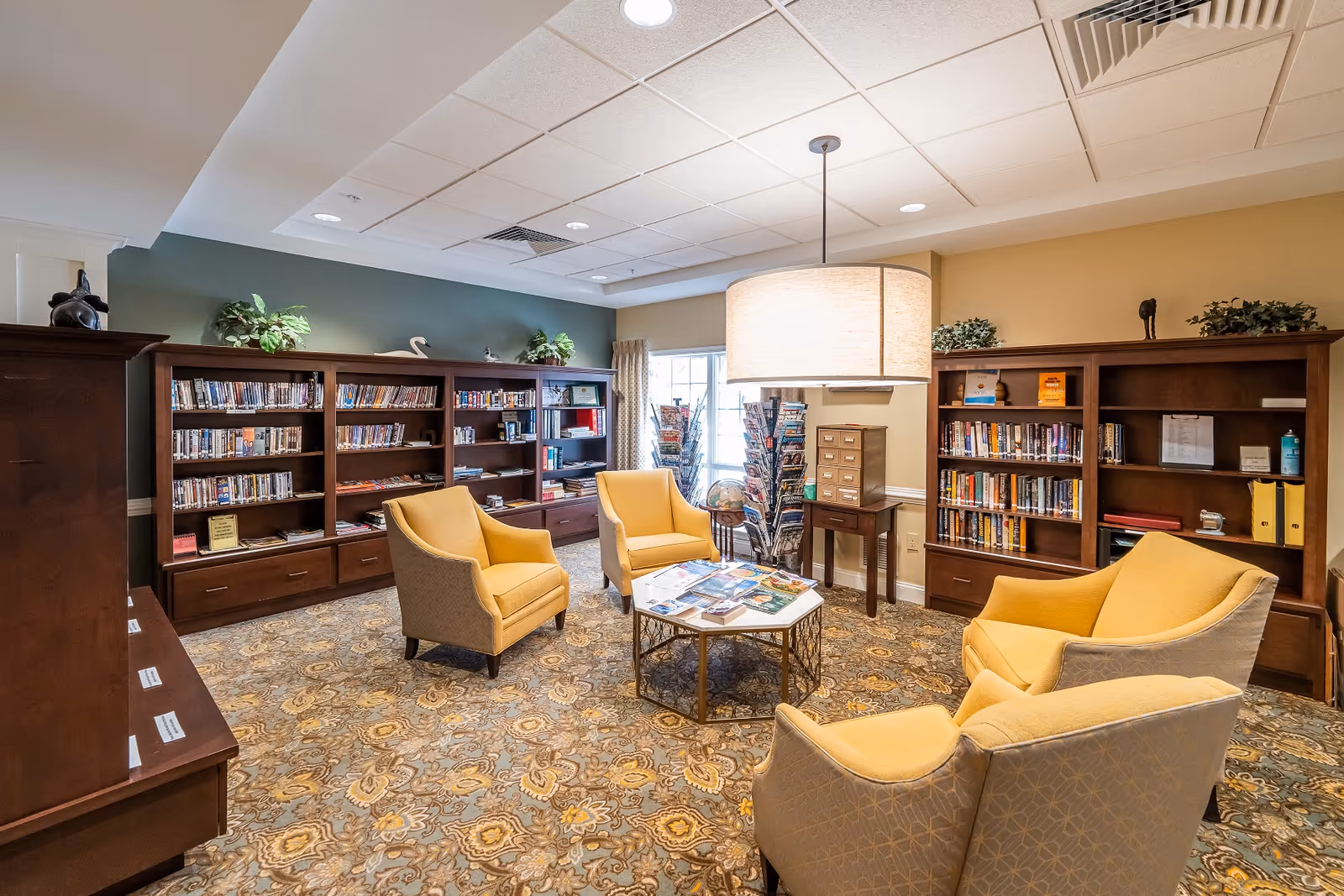 Cozy senior living community library/lounge with bookshelves, yellow armchairs, a central table and overhead pendant light.