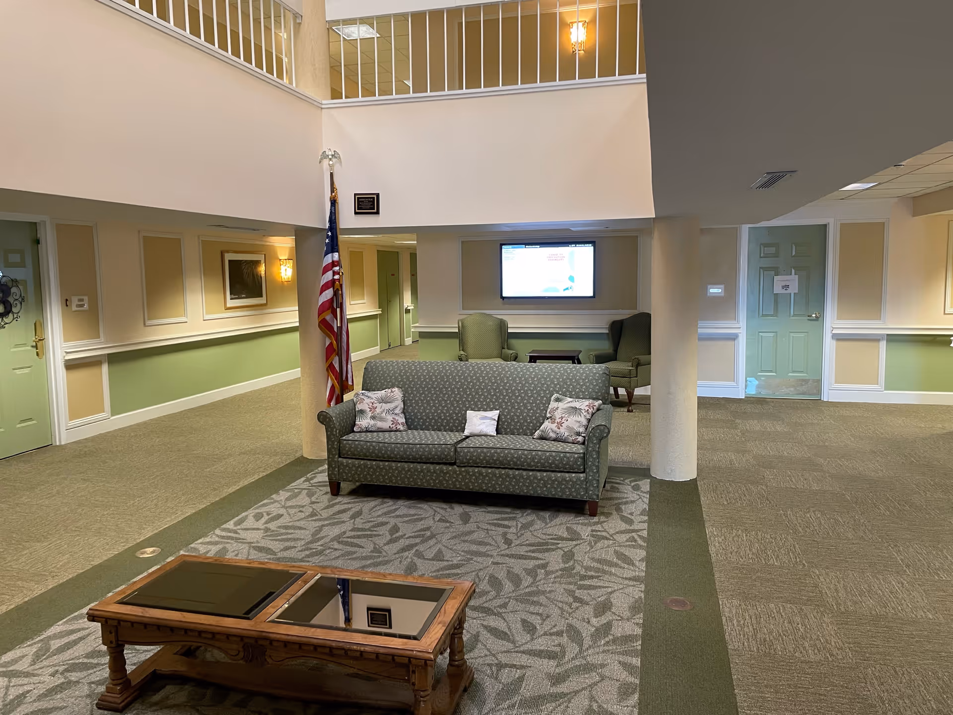Interior view of a senior living facility lounge area with a green patterned sofa and two floral pillows, a wooden coffee table with glass inserts, two green armchairs in the background, an American flag, and a wall-mounted screen. The walls are painted beige and green with white trim, and there is a green door on the left and another door in the background.