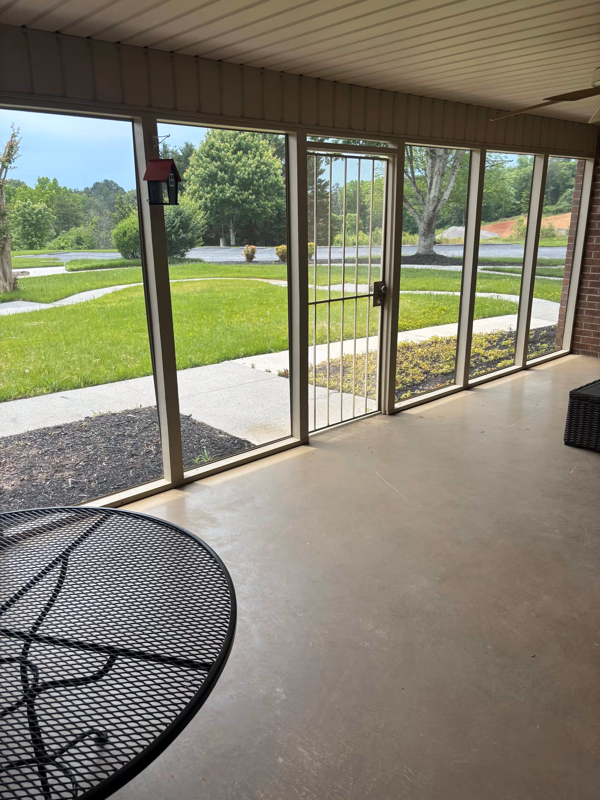View from a covered patio area with a metal mesh table in the foreground. The patio is enclosed with large screened windows and a metal security door, looking out onto a green lawn, trees, and a paved walkway.