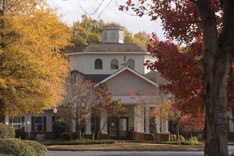 Front exterior view of a senior living facility building surrounded by trees with autumn foliage in shades of yellow, orange, and red. The building has a central entrance with columns and a cupola on the roof.