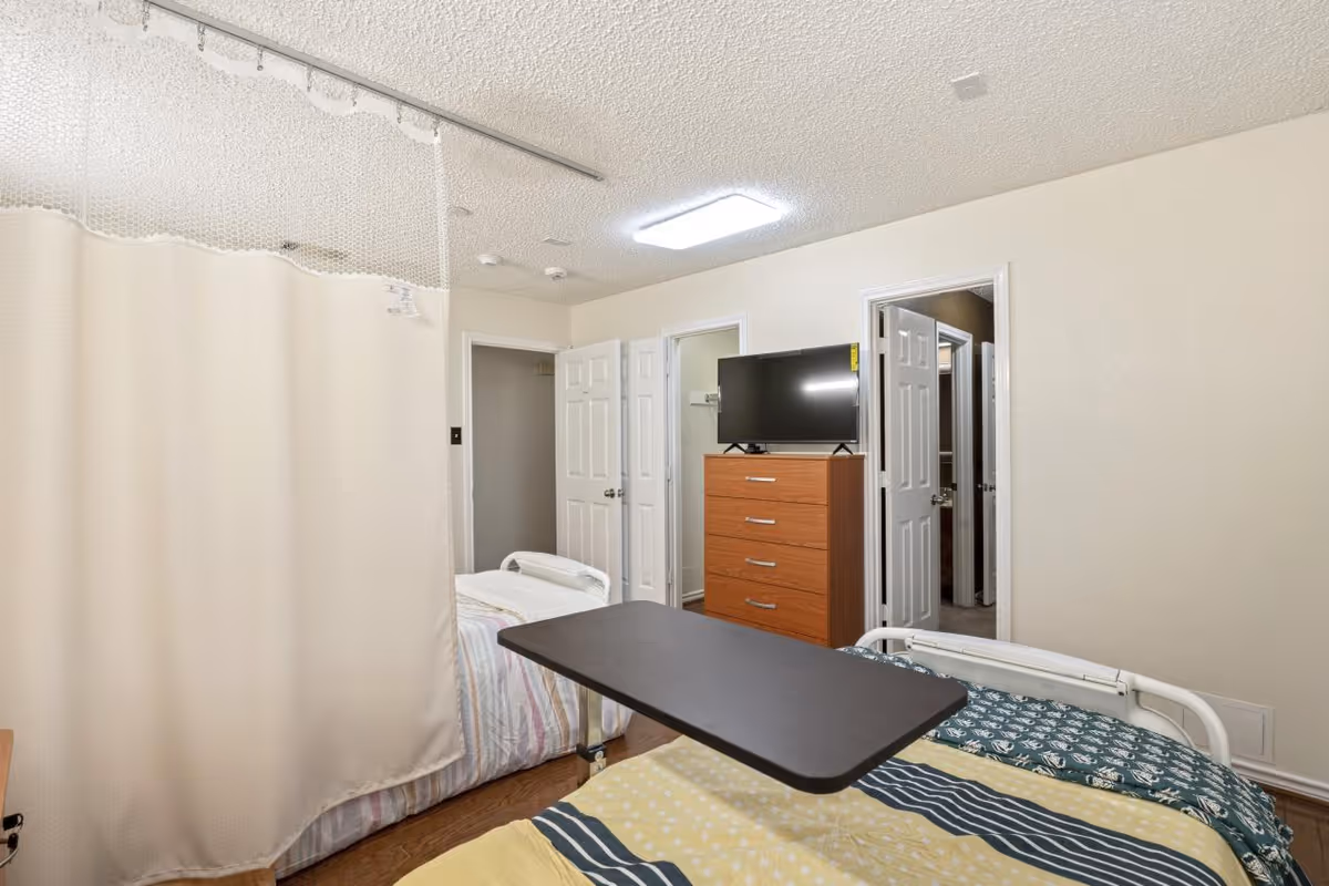 A shared assisted living bedroom with two beds separated by a privacy curtain. Each bed has a different patterned bedspread. There is a wooden dresser with a flat-screen TV on top, and two open doors leading to other rooms. The room has light-colored walls and a ceiling light.