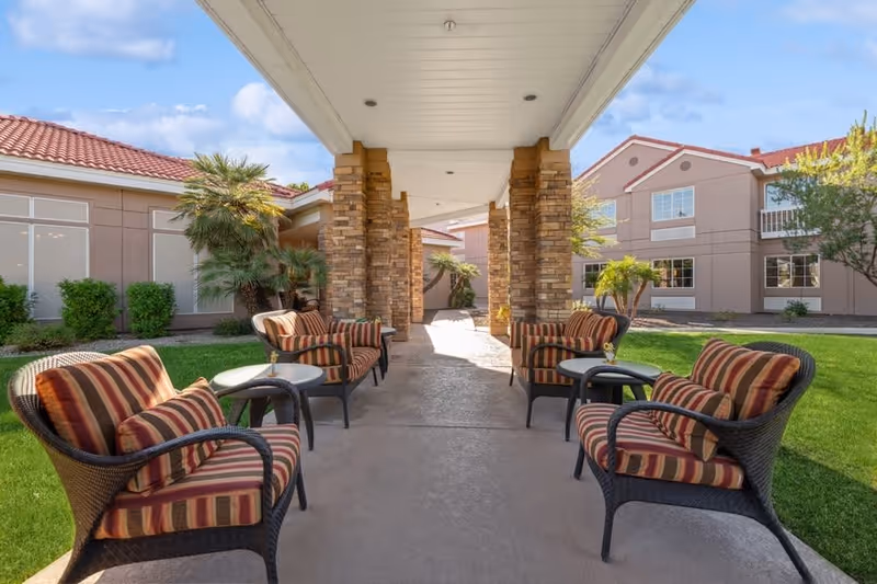 Outdoor covered seating area with six cushioned wicker chairs arranged around three small round tables. The area is flanked by stone pillars and surrounded by green grass, palm trees, and beige buildings with red tile roofs under a partly cloudy blue sky.