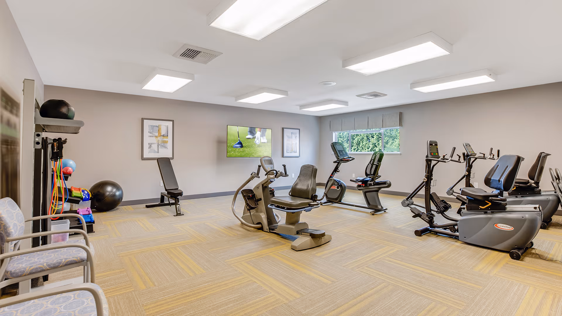 A well-lit fitness room with exercise equipment including recumbent bikes, an adjustable bench, resistance bands, and exercise balls. There are two chairs on the left side and a wall-mounted TV displaying a golf scene. A window on the right wall lets in natural light.