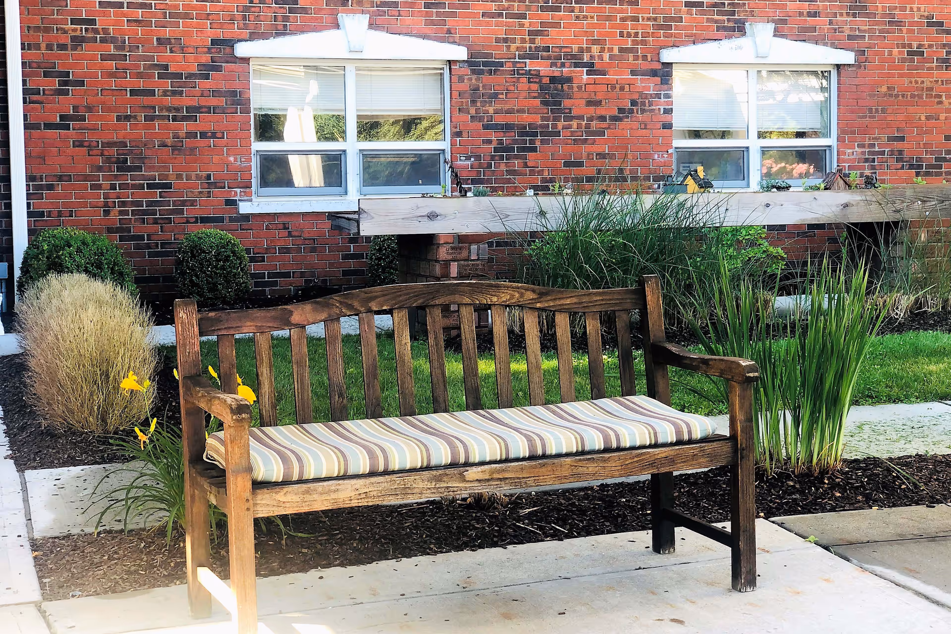 Wooden bench with a striped cushion in a landscaped courtyard in front of a red brick building with windows.