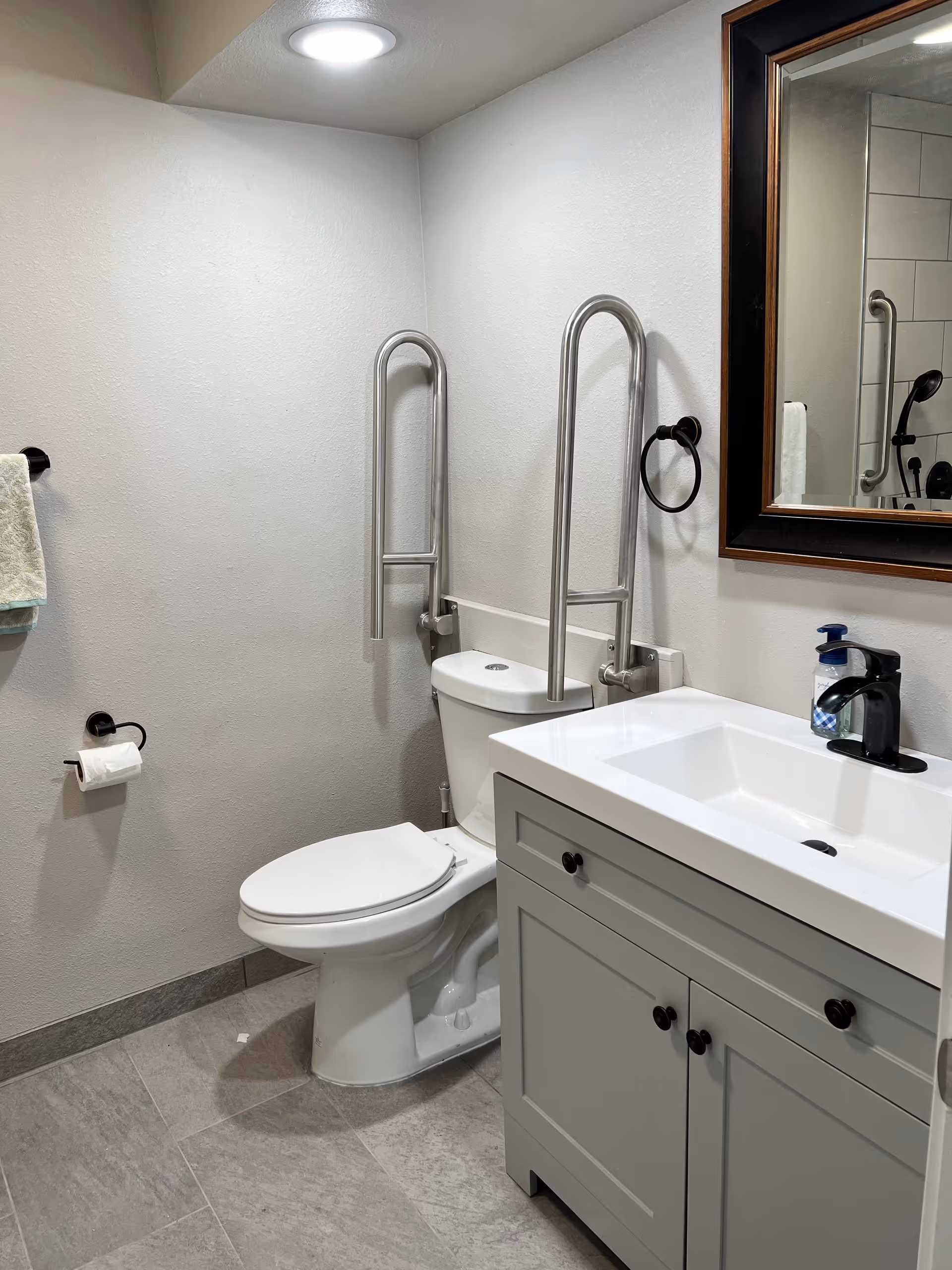 Bathroom with a toilet fitted with stainless steel grab bars, a sink vanity with mirror, towel ring and soap dispenser.