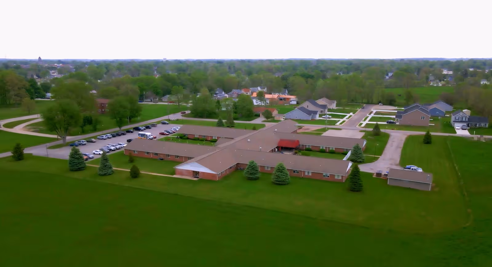 Aerial view of Clinton House Rehabilitation and Healthcare Center, a single-story building with a brown roof surrounded by green lawns and trees. There are several cars parked in the parking lot adjacent to the building, and residential houses are visible in the background.