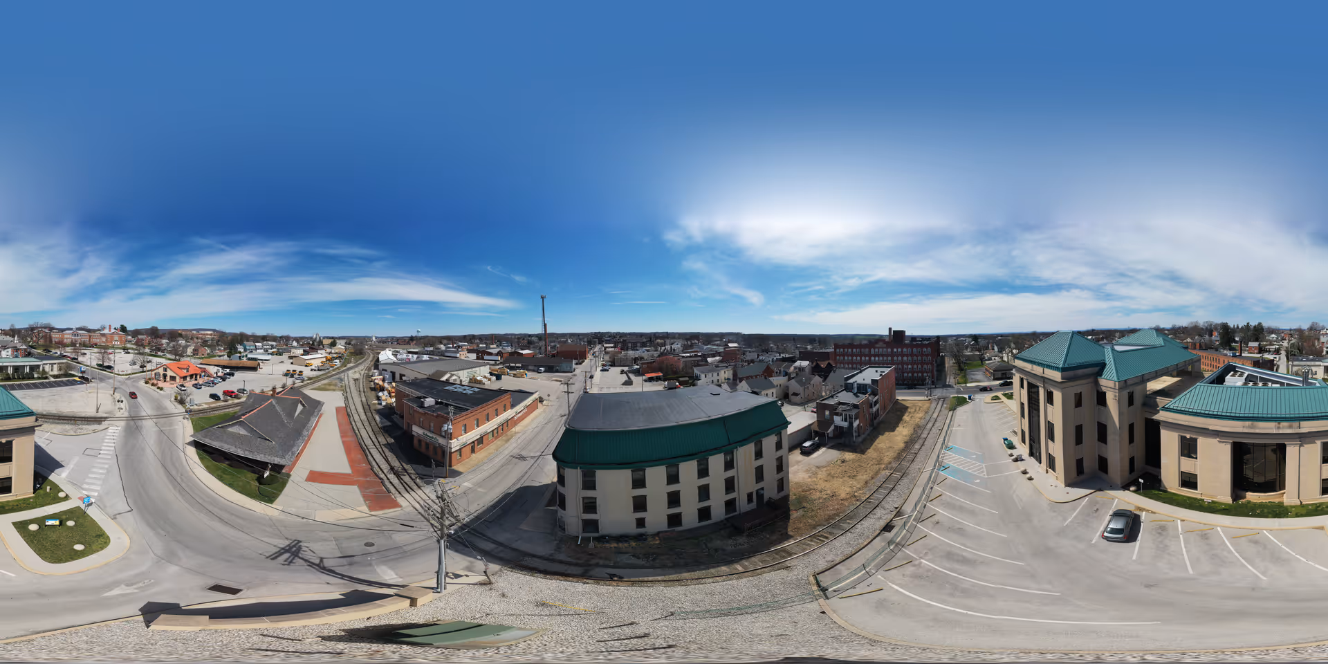 Aerial panoramic view of a small town center showing roads, railroad tracks, parking lots, and several buildings with green roofs under a blue sky.